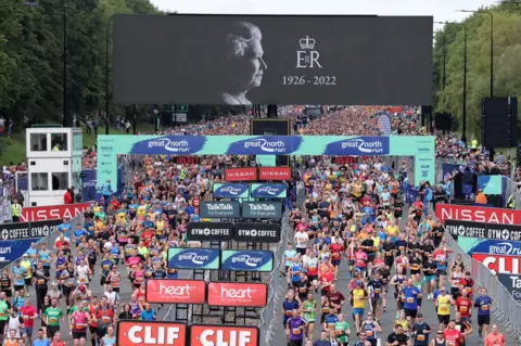 Raoul Dixon/NNP Wide view of hundreds of runners starting the run in front of a large video screen showing the Queen