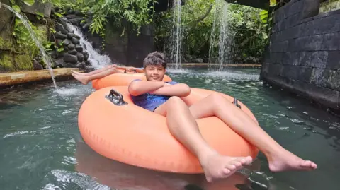 A young boy, with his arms crossed, floating on a inflatable orange ring in a pool with greenery and a waterfall in the background 