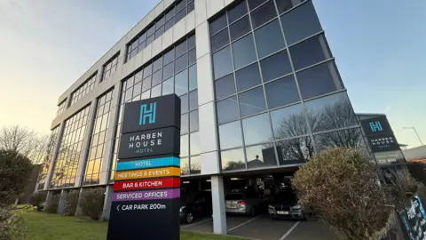 STEVE HUBBARD/BBC Exterior shot of Harben House Hotel, a mainly glass angular building with a sign in front of it. In front of a blue sky. There is bush to the right of the picture.