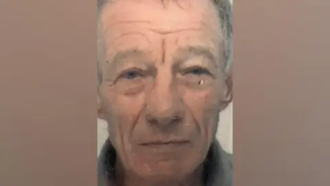 LancashireConstabulary Brian Fletcher stares forward in a dated photo released by police. He is wearing a grey shirt and has grey hair with a scar below his eye.