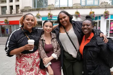 Four women, standing with their arms round each other in front of a Poundland shop. One is holding a hot drinks cup.