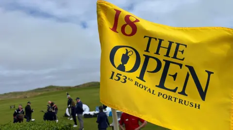 A yellow golf flag on the Royal Portrush course with 'The Open 153rd Royal Portrush' written in navy letters