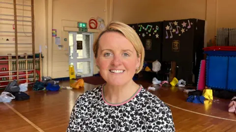 A woman with short blonde hair is standing in a school hall. There are different coloured schoolbags behind her on the wooden floor.