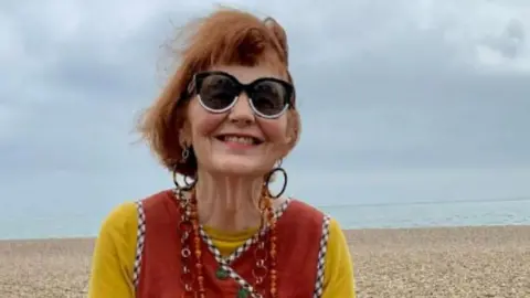 Marcia Riddington with red hair sits on a sea wall smiling at the camera. She wearing a yellow top with a red vest over the top and a skirt made up of a former quilt with different patches of fabric.