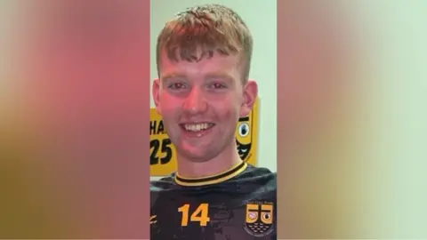 PSNI Pádraig McDonald. He has short ginger hair, wearing a black and yellow football top.