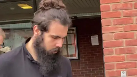 Dylan McNamara leaving court. He is wearing a black polo shirt and has long brown hair tied in a bun and a long black beard.