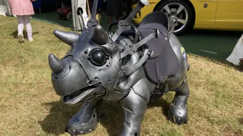 A robotic looking dinosaur stands in a grass field. It is a dark grey colour and has three horns on its head. It has a black saddle fitted to its body.