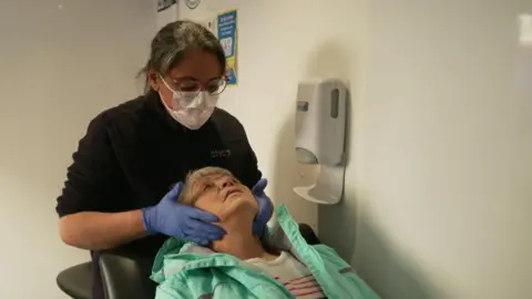 A dentist with glasses is wearing a fleece and a white face mask as well as blue disposable blue gloves. Another woman, wearing a light-green jacket and white jumper with red stripes, is lying back on a black chair. The dentist is using her hands to feel the woman's neck and throat.