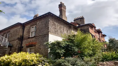 Richard Fisher College House, on the campus of the University of Leicester. The old house is pictured in the sunshine. There are scattered clouds. It is surrounded by shrubs and trees. 