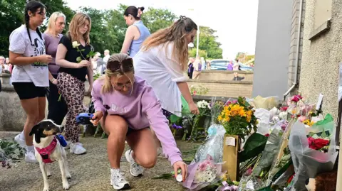 A woman placing a candle on the ground beside a pile of flowers. There are five women behind her and a dog beside her. 