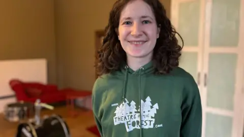 Jon Wright/BBC Rei Modue, wearing a green 'Theatre in the Forest' hoodie, stands in a rehearsal room. There is a drum kit in the background.