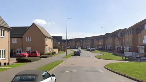 A street in a housing estate. It has two-storey yellow brick houses on the left and three-storey yellow brick houses on the right. There are a few parked cars on the street and grass verges.