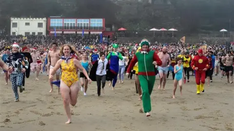 Getty Images Crowds of people dressed as superheroes run towards the camera grinning down the beach. 