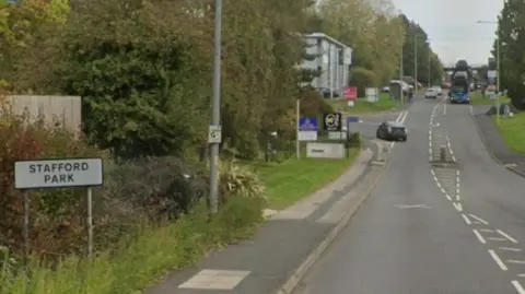 Google A white road sign that reads "Stafford Park" on the side of a road. The road is has two single carriageways and goes off into the distance. There is a junction of the left which a car is turning into. A bus, which is driving towards you, is approaching in the distance.
