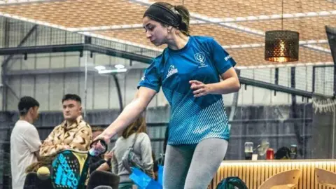 Daisy Whitly, uma jovem, vestindo uma campanha e uma equipe esportiva azul está em direção à bola de chegada. O tiro do público é quase moinho ao fundo, que parece ser levado para dentro de um centro de aposentadoria com paredes de concreto alto.