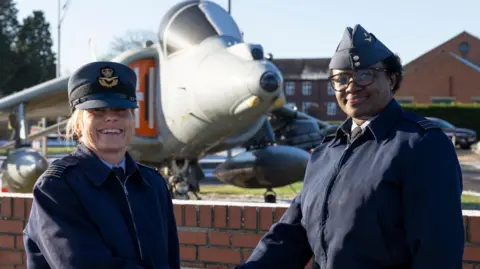 RAF Wittering Wing Cdr Nikki Duncan (left) and Wing Cdr Joan Ochuodho are both dressed in blue navy uniform jackets and hats. They are smiling at the camera and appear to be shaking hands. (Their hands are out of shot.) They are standing in front of an aeroplane.
