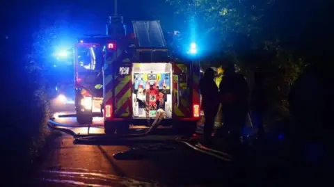 Fire engines parked on a road at night.  Blue lights and hoses coming from the rear can be seen and a group of firefighters are to the right.