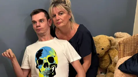 Hazel Howe Thomas Howe with short dark hair and a white T-shirt with a skull motif on the front. There is a brown teddy bear behind him, sitting alongside a blue wall.