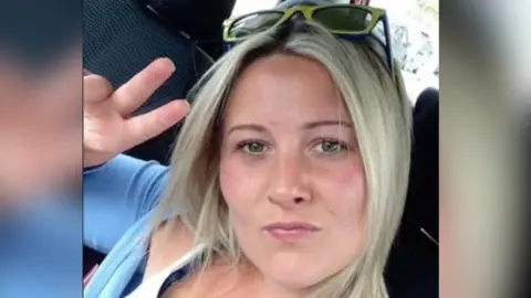 Joanne Penney seen in selfie taken sitting in a car. She is making the peace sign hand gesture and has shoulder-length straight blonde hair and wears a light blue cardigan.