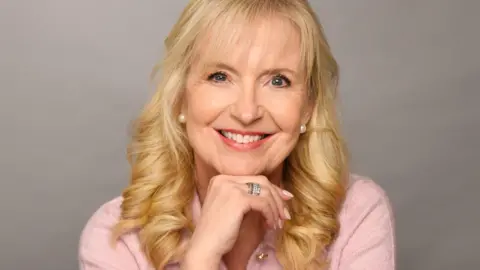 Carol Kirkwood smiling and resting her chin on her hand while wearing a pink top