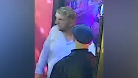 Blurry CCTV image of two middle aged men - one is wearing a black jacket and flat cap with his back to the camera, and the other is facing towards the camera, with shaggy but short greying blond hair and a blue long-sleeved shirt with the top button undone.