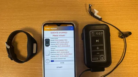 A watch, smartphone and the portable device with an ear piece on a table. The smartphone displays an interface to control the device, saying "service stopped" and "press 'start".