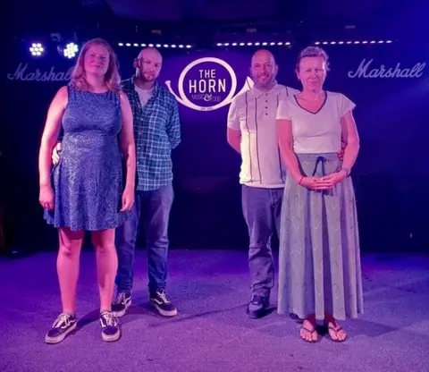 Luke Hinton Four people standing together on a stage, illuminated with purple lights