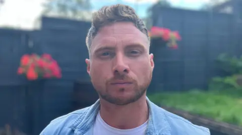 Sam Read/BBC Craig Spokes is looking at the camera with a serious expression and wearing a white T-shirt with a blue shirt open on top. He is standing in a garden with hanging baskets in the background.