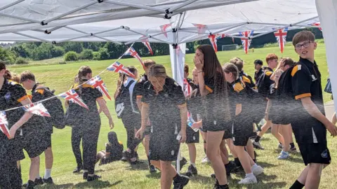 Beacon Hill Academy Pupils in dark coloured sportswear walk through a white gazebo while sprinklers spray them with water.