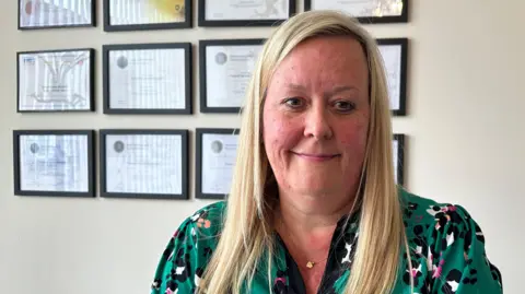 BBC/Matt Dean A smiling woman with long blond hair wearing a green patterned dress. There are twelve black-framed certificates behind her on a beige wall.