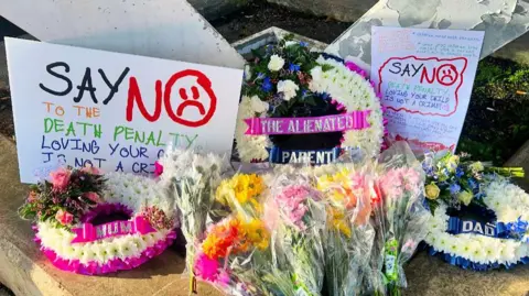 Simon Cobb There is a funeral wreath at the centre of the picture with the words "the alienated parent" written on it and posters either side saying 'No to the Death penalty". There are two other smaller wreaths, one with the word 'Mum' the other with the word 'Dad' written on a ribbon. There are several bunches of flowers in the foreground. 