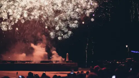 St Ives Business Improvement District Fireworks explode above a pir with a lighthouse at the end . In the foreground are crowds of people watching the display 