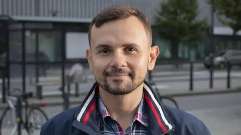 Dalibor Sentic Dalibor Sentic is photographed stood outside on a street. A bike, bus stop and trees can be seen behind him. He has brown eyes, short brown hair, and a beard. He is wearing a plaid grey, red and white shirt and navy-blue jacket, which has a white and red stripe on the collar.