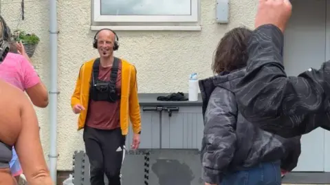 A male fitness instructor wearing headphones and a yellow cardigan is facing the camera, dancing with a group of women whose backs are towards the camera, some with their arms in the air