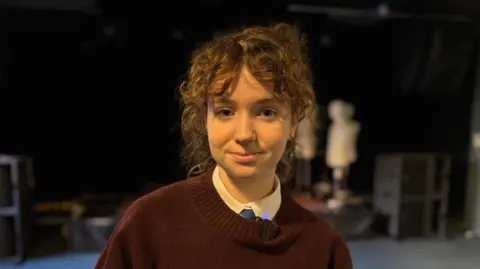 Headshot of 15 year old girl with red hair. She is standing in the event space in front of a stage with mannequins. She is wearing a shirt and blue tie with a brown jumper over it.