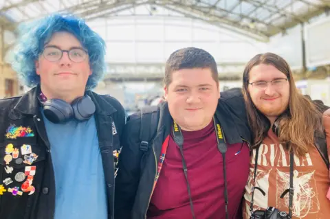 Three young men - Danny, Kieran and Liam - smiling at camera in a train station, all wearing bright t-shirts and carrying cameras.