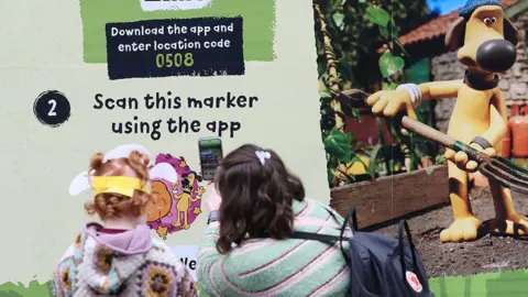 Liverpool ONE A woman with shoulder length brown hair with a grey pink and red striped jumper with a young girl at the side of her with orange curly hair wearing a multi-coloured knitted cardigan points. The woman points her mobile phone at a QR code of Shaun the Sheep app trail on an exhibition board which also features a picture of Bitzer the dog from CBBC's Shaun the Sheep.