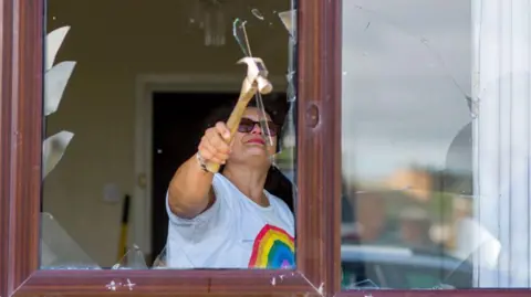 Getty Images A pistillate   wearing a achromatic  T-shirt with a rainbow connected  the beforehand   stands wrong  a house. In beforehand   of her is simply a breached  model   pane. She is holding up   a hammer and breaking the remaining bits of solid  retired  of the pane.