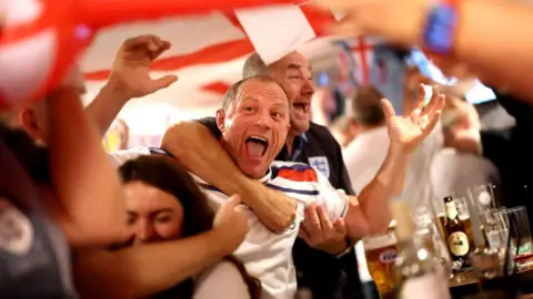 Getty Images Two male football fans wearing England shirts celebrate a goal in the final of Euro 2024 - one of the men has his arm around the other, and are screaming in celebration 