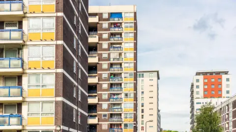 A side view of three older residential tower blocks in a row with a a new block to the right of the image. 