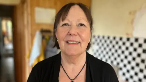 A woman with dark hair in a short bob is looking into the camera and smiling. She is wearing a black cardigan and black pendant necklace. 