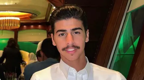 Naif Alqassim Mohammed Algasim has black hair combed back and a moustache. He is wearing a white shirt and smiling while sitting at a restaurant table.
