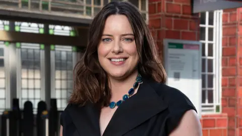 Michelle Ryan is smiling on the set of EastEnders, she is stood in front of Walford tube station