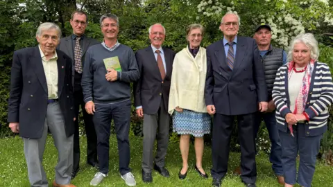 A row of people standing in the garden, there are six men and two women. One of the men is holding a copy of the book titled "The Good Doctor".