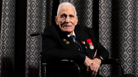 PA D-Day veteran John Dennett, from Liverpool. He is wearing a dark suit with medals on it and sitting in a wheelchair. The background is geometric grey curtains. 
