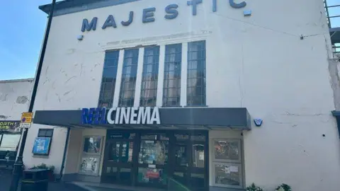 The front of a large white, block shaped building with black-framed doors and a sign above the door which says Reel Cinema