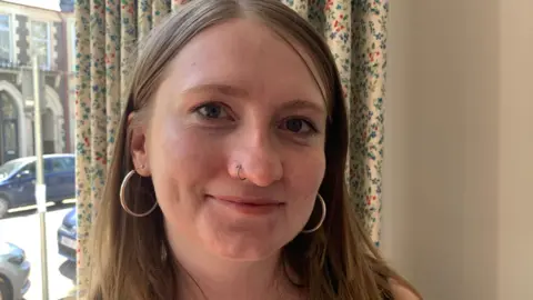 Marianne looks at the camera, she's wearing hoop earrings and has a nose piercing. She's smiling and a curtain and window overlooking the street is visible behind her. 
