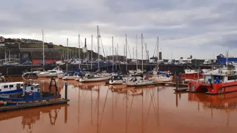 BBC Weather watchers / Gadabout About two-dozen boats are parked up in the harbour. They are connected by a wooden jetty and stairs lead up to the carpark beside it. The water is dark orange.