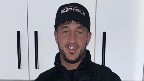 A photograph of Kaine Fletcher, a mixed-race man in his twenties, weather a black cap which reads 'KJ Mobile' and a black jacket. He is standing in front of white cabinets. 