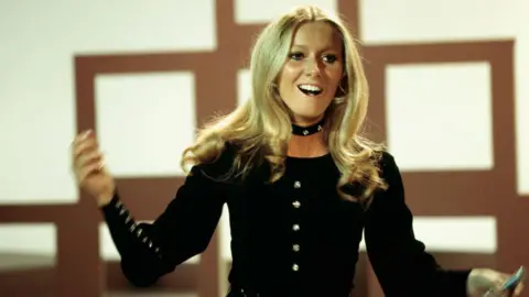 Getty Images Clodagh is photographed singing on stage and she has her arms raised from her sides. She has long blonde hair and is wearing a long sleeved black top that has silver buttons down the middles. She is wearing a black choker style necklace. 
